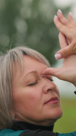 de mediana edad con las manos retorcidas y tocando su frente, ojos cerrados, practicando yoga al aire libre en un campo de hierba brumosa, rodeada de árboles, material verde y amarillo visible en un fondo borroso