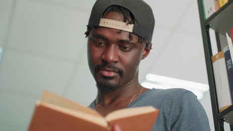 Close up of young man wearing cap reading and flipping through brown hardcover book in library, with bright ceiling lights and bookshelf in background
