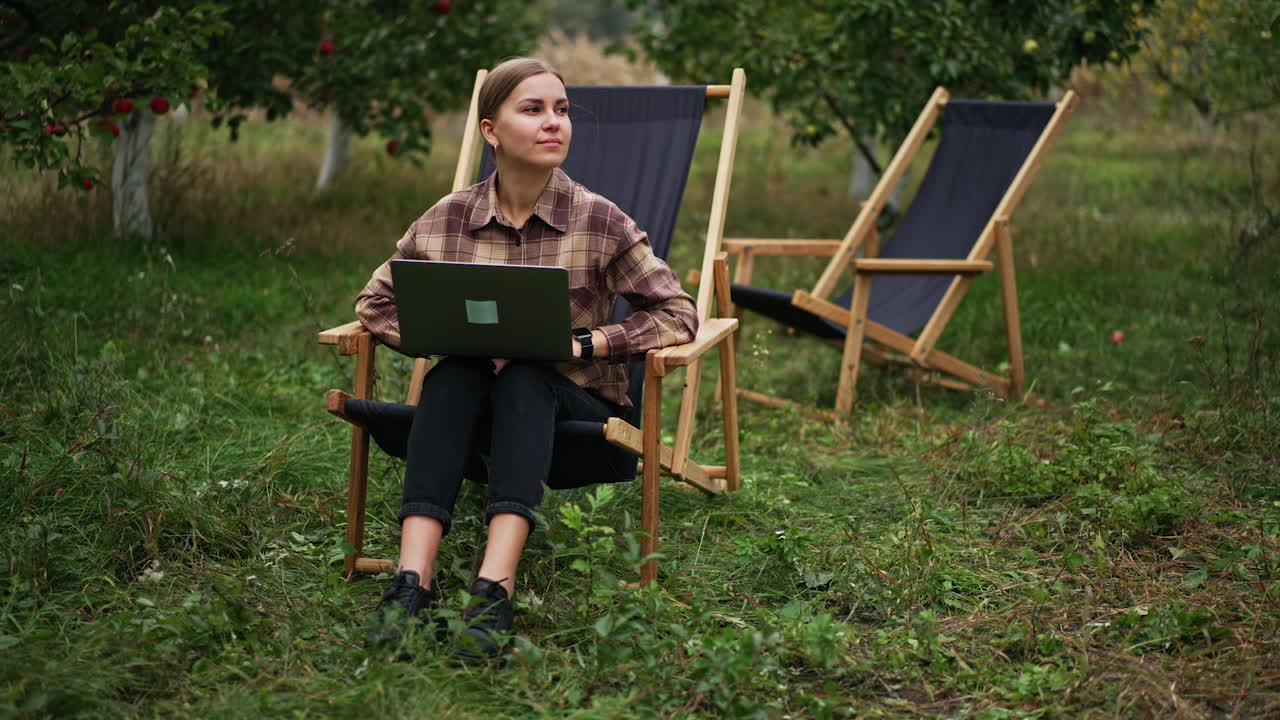 Pensive lady in plaid shirt sitting in garden chair working on laptop. Freelance working woman thinking over some work issues. Nature backdrop.