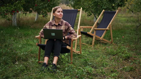 pensive lady in plaid shirt sitting in garden chair working on laptop. freelance working woman thinking over some work issues. nature backdrop.