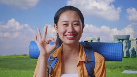 Close Up Of Asian Female Hiker With Mountaineering Backpack Smiling And Showing Okay Gesture To Camera While Traveling In Stonehenge