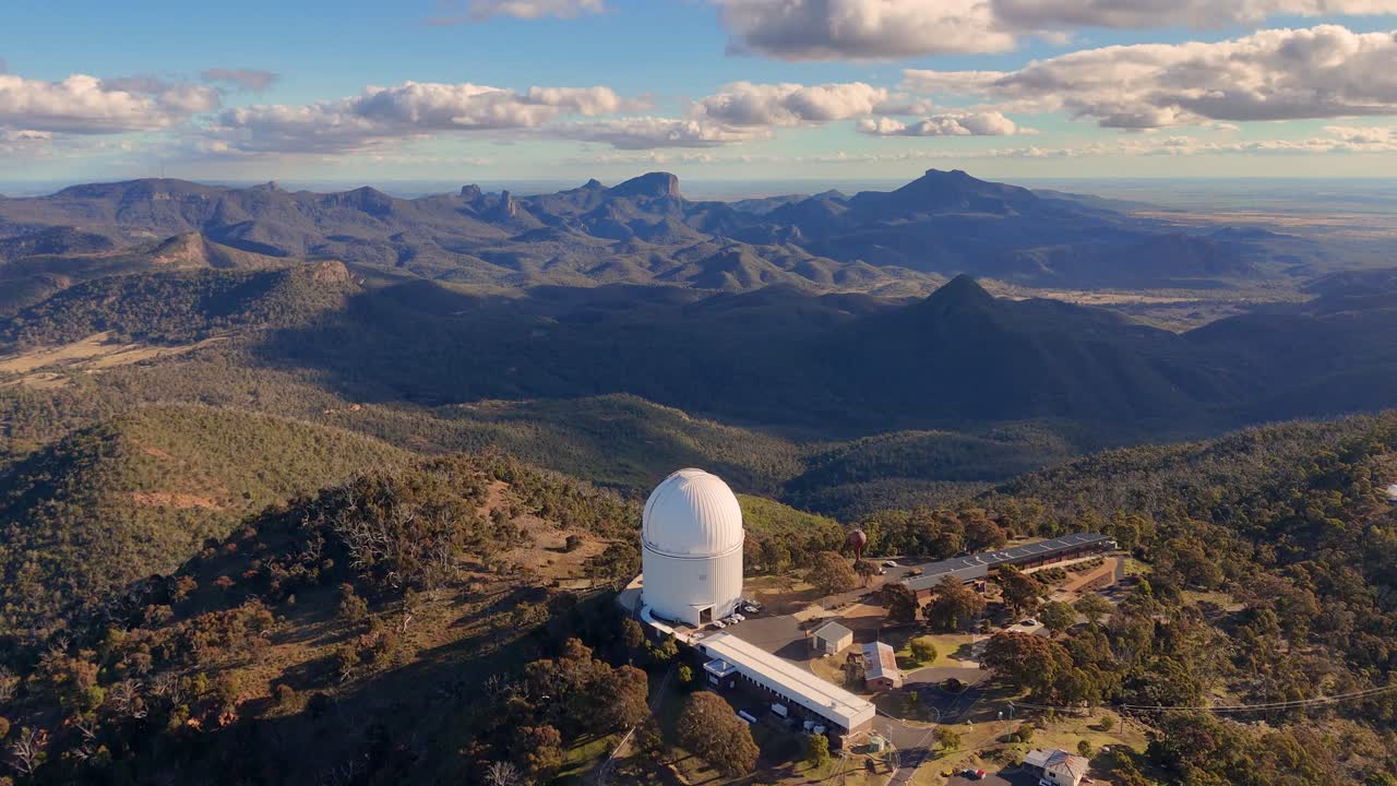 Aerial drone footage orbits a large white observatory dome atop a forested mountain ridge in Australia, with dramatic late afternoon sunlight and expansive views