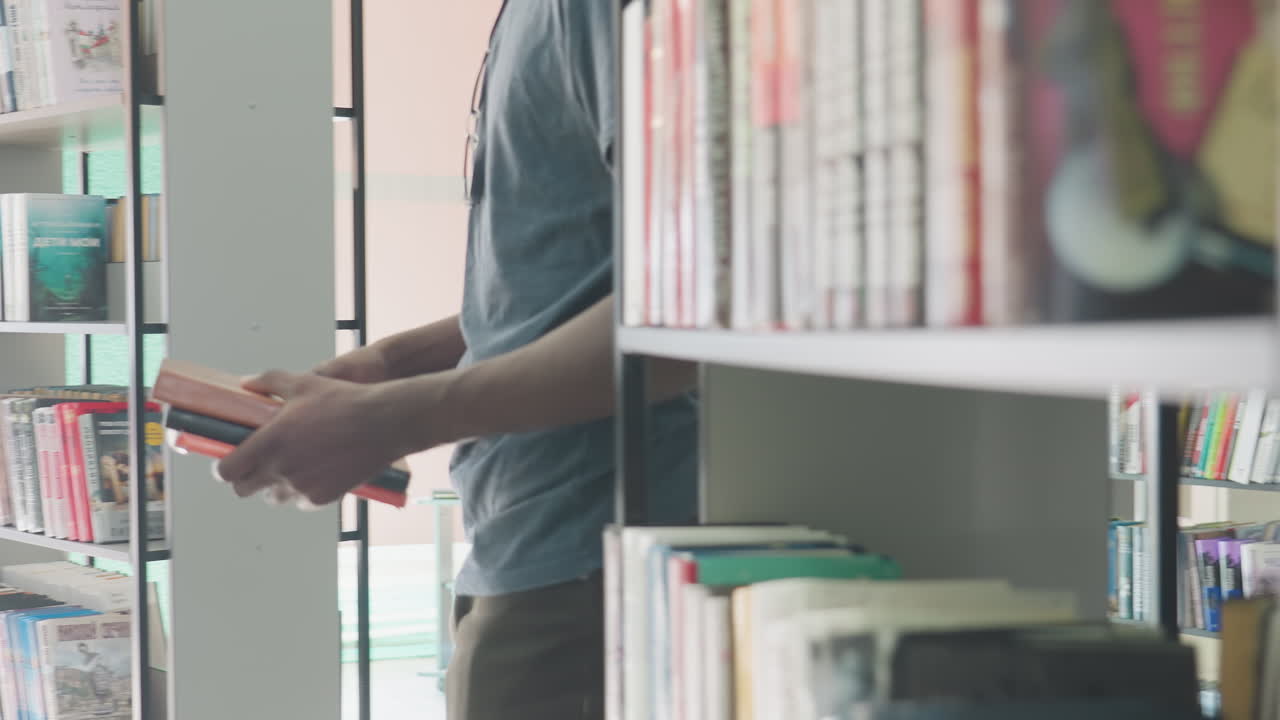 Primo piano di una persona che tiene in mano diversi libri all'interno di una luminosa biblioteca moderna, mentre seleziona un altro libro dallo scaffale circondata da dorsi colorati e letteratura organizzata sullo sfondo