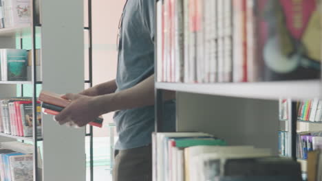 close up of person holding multiple books in hands inside bright modern library, selecting another book from shelf while surrounded by colorful spines and organized literature in background