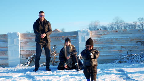 caucasian family of three is outdoors in winter on sunny day. adorable toddler boy picks some snow and walks approaching camera.