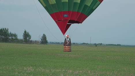 rear view of passenger riding in colorful hot air balloon gliding low over vast green farm land at sunset with burner flame flickering against pastel sky while distant trees and fence line