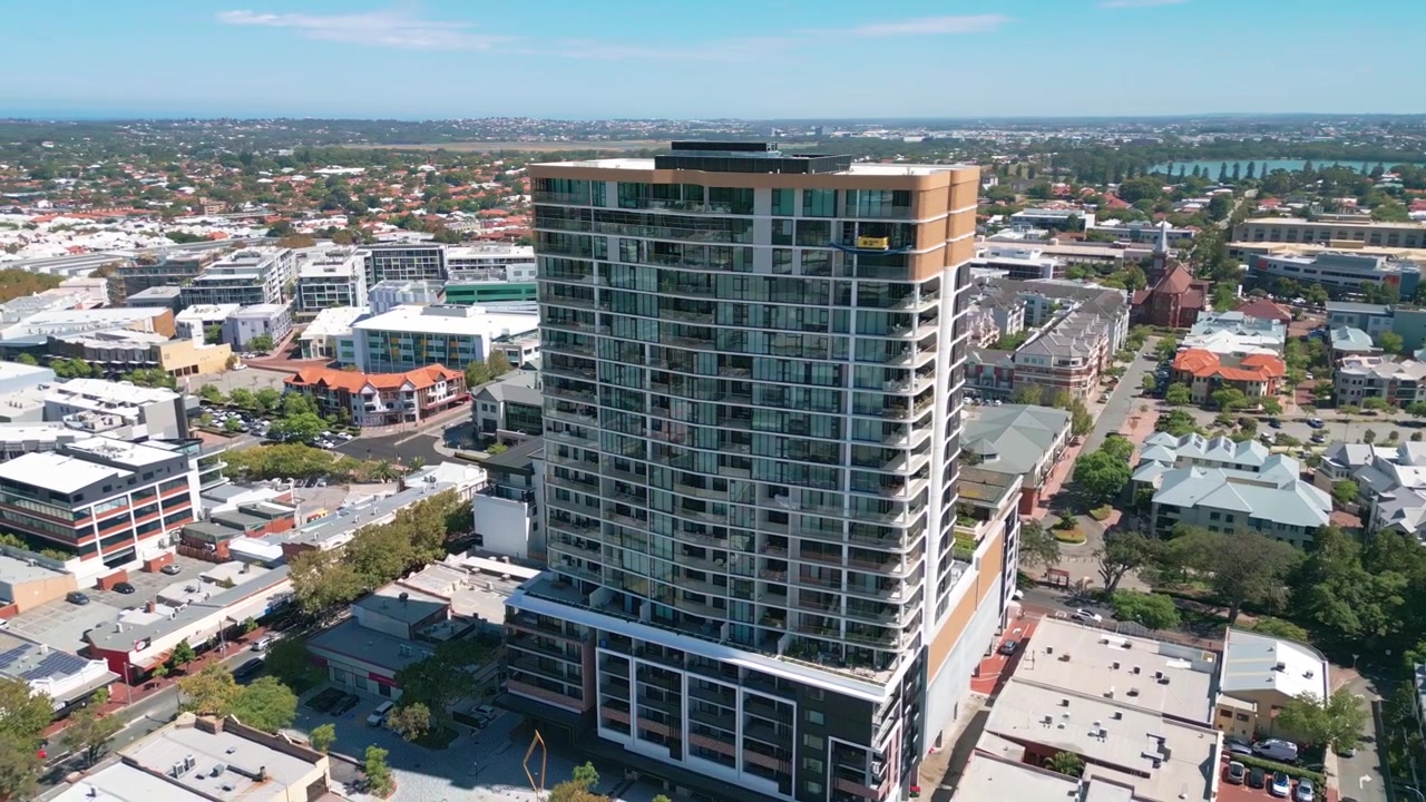 Tomada orbital em torno de uma moderna torre residencial de vidro de arranha-céus, revelando a faixa comercial de Subiaco, igreja histórica, ruas suburbanas e um rio distante ao fundo sob céu azul. Subiaco, Perth, Austrália