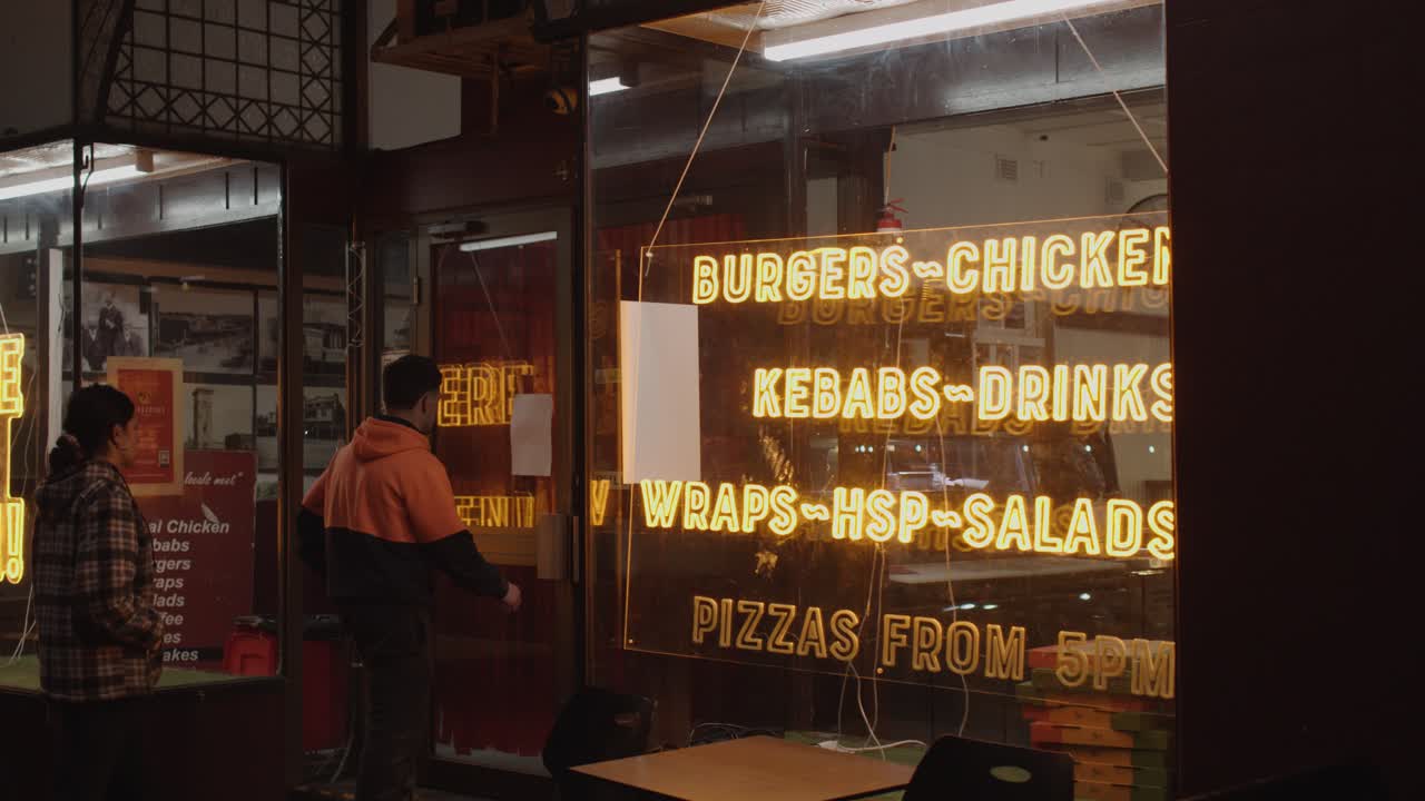 Two people approach and enter a brightly lit takeaway shop under neon signage at night