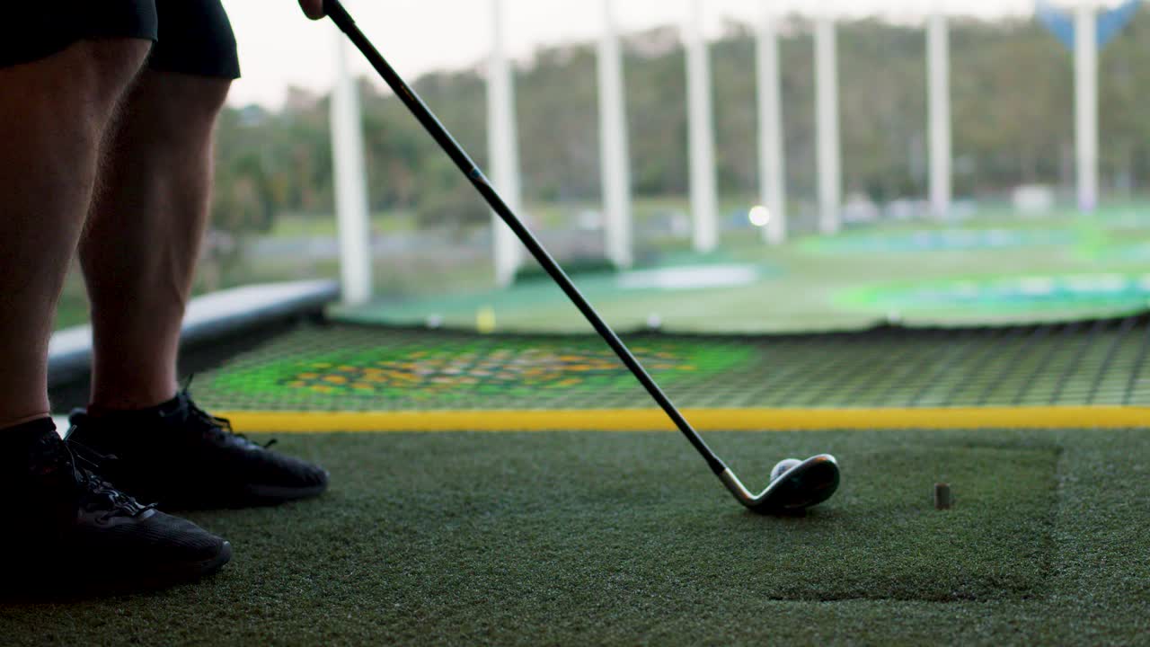 Golfer hits ball on artificial turf at driving range, low angle, natural daylight, steady shot