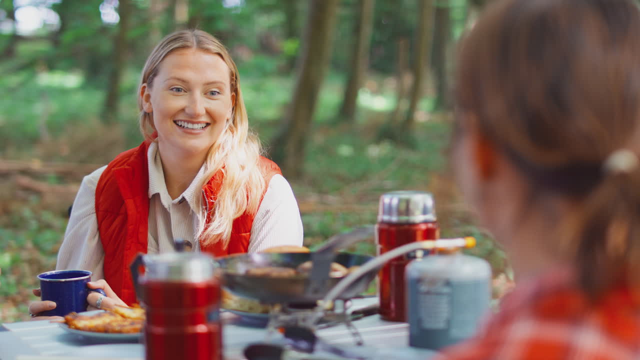 두 명의 여성 친구가 함께 텐트에 앉아 숲에서 요리하는 휴가에 있습니다.