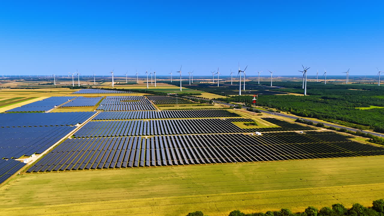 Mowed agricultural fields with installed solar panels. Modern wind turbines work at backdrop. Aerial view
