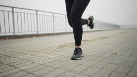 leg view of sports person jogging in black sneakers and leggings on outdoor pathway near railing, shoelace swaying during movement, with misty background and focus on dynamic motion