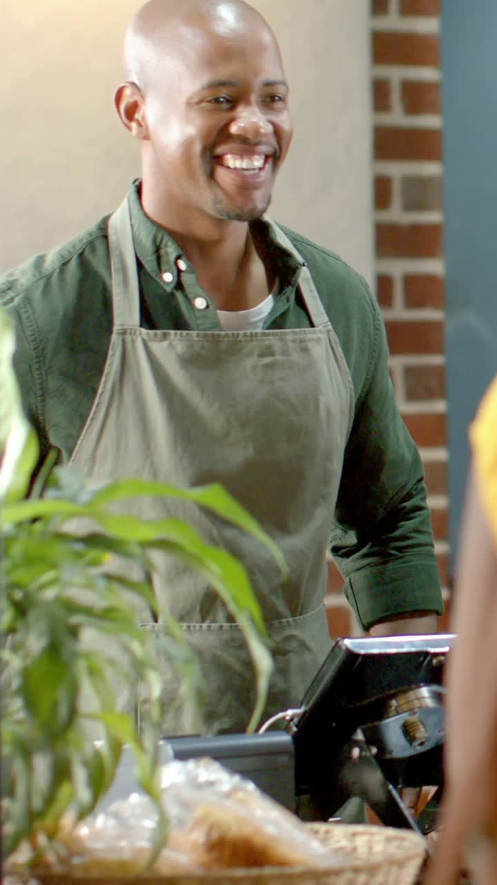 Vertical video: Smiling worker in apron serving customer entering right at cafe counter with till