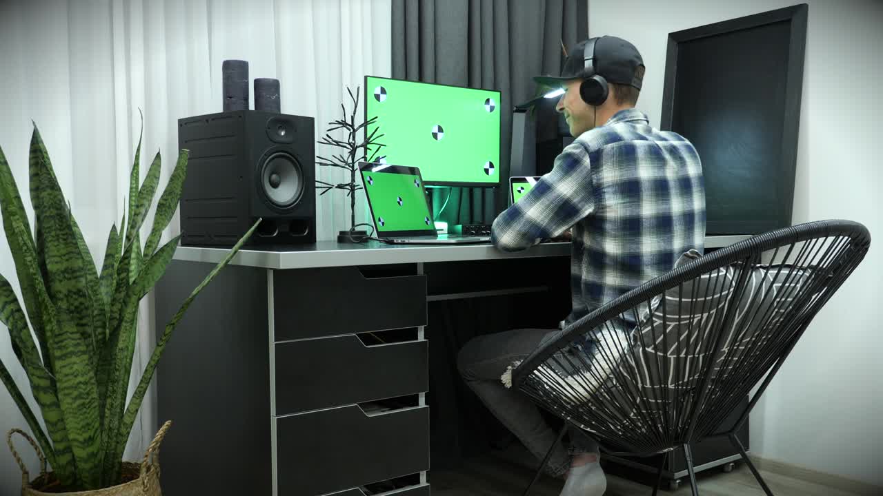 홈 오피스에서 녹색 크로마 키 스크린을 가진 노트북을 사용하는 남자, 집에서 일하는 젊은 남성 프리랜서, 키보드에서 타이핑, 헤드폰에서 음악을 듣는 비즈니스 개념