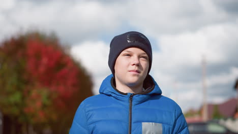 young boy in blue jacket and black beanie strolls in residential area, looking thoughtful with sunlight reflecting off his face against blurred autumn trees and houses