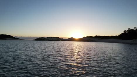 Antena-Baja-Sobre-El-Agua-Hacia-El-Amanecer-Revelando-Olas-Rompiendo-En-Una-Playa