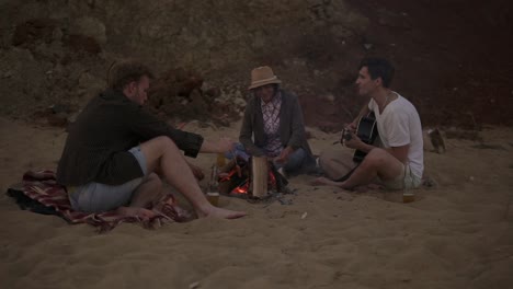 amigos jóvenes sentados junto al fuego en la playa por la noche, asando salchichas, bebiendo cerveza y tocando la guitarra. cámara lenta