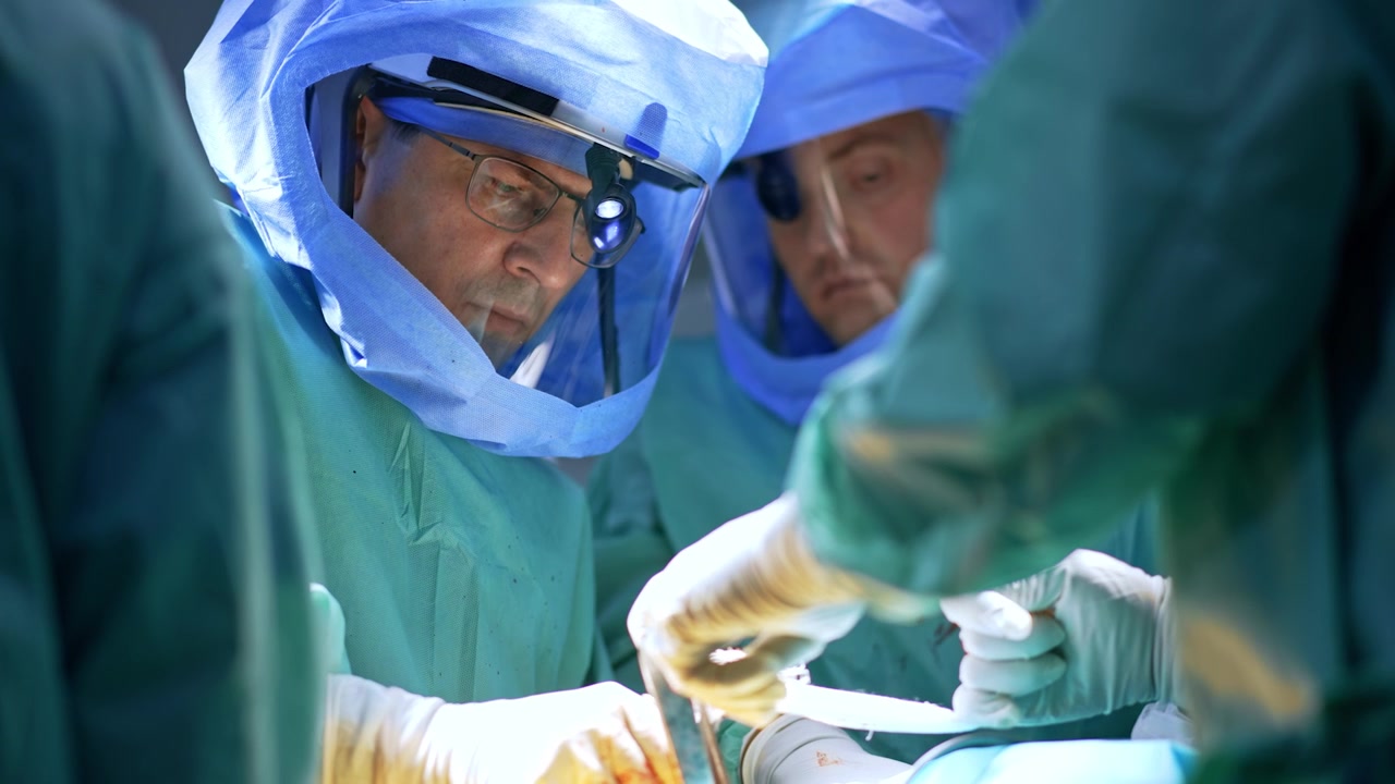 Confident male Caucasian surgeon in glasses and protective helmet performs surgery assisted by the group of medics. Focused doctor applies medical tools.