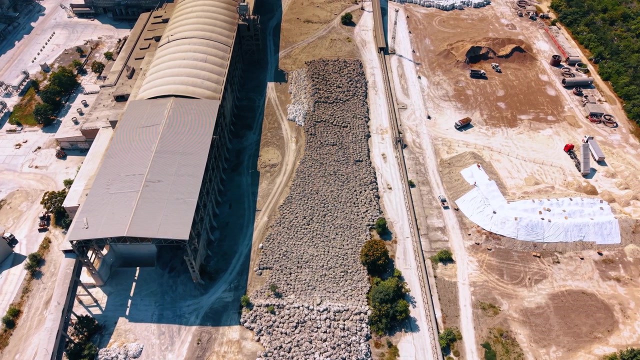 High angle view of a factory warehouse and raw materials.. A long building sits next to a large stone pile at an industrial facility.