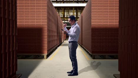 full body side view of an asian male professional worker standing with his phone with red brick packed in stacks are stored, he raises his fist up with screaming goal