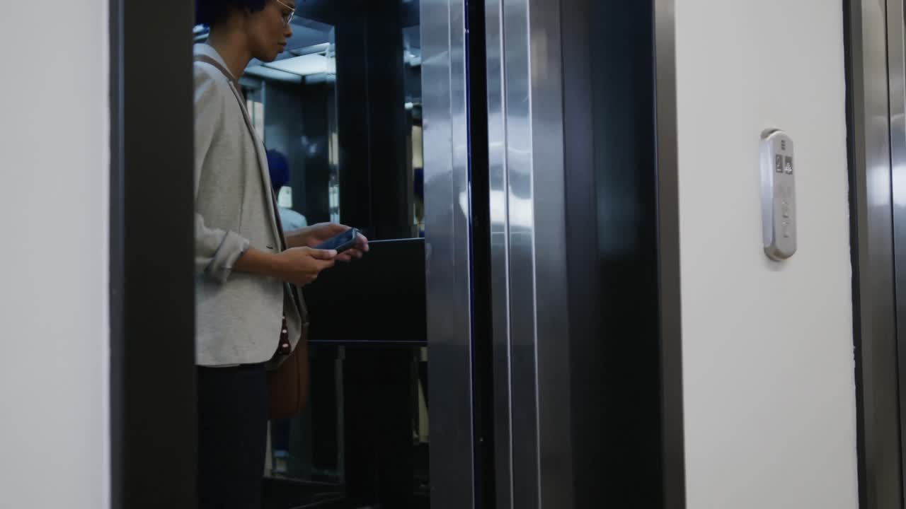 mujer de negocios biracial con afro azul usando un teléfono inteligente y entrando en el ascensor en la oficina, cámara lenta