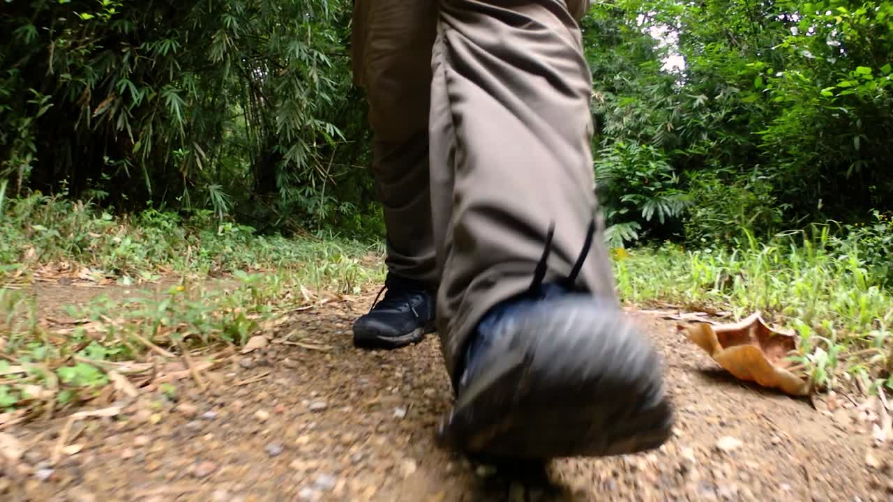 un hombre caminando en la jungla, la cámara enfocada en sus piernas, mostrándolo dando un paso adelante en el camino de tierra con sus zapatos enmarcados