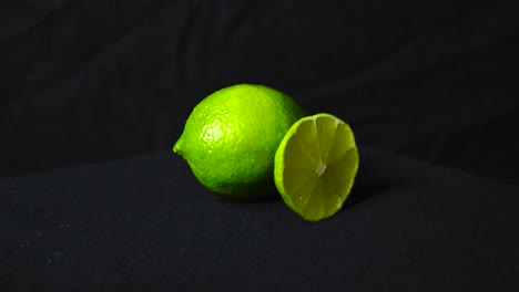 footage zooming or closing in on two vibrant green lime fruit. one is cut in half and the other is whole. the healthy fruit have been placed on a black dark display with a black background under light