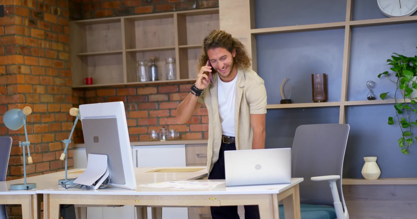 Uomo che riceve una chiamata, tiene il telefono, guarda il laptop e i documenti sulla scrivania in ufficio, discutendo di lavoro.