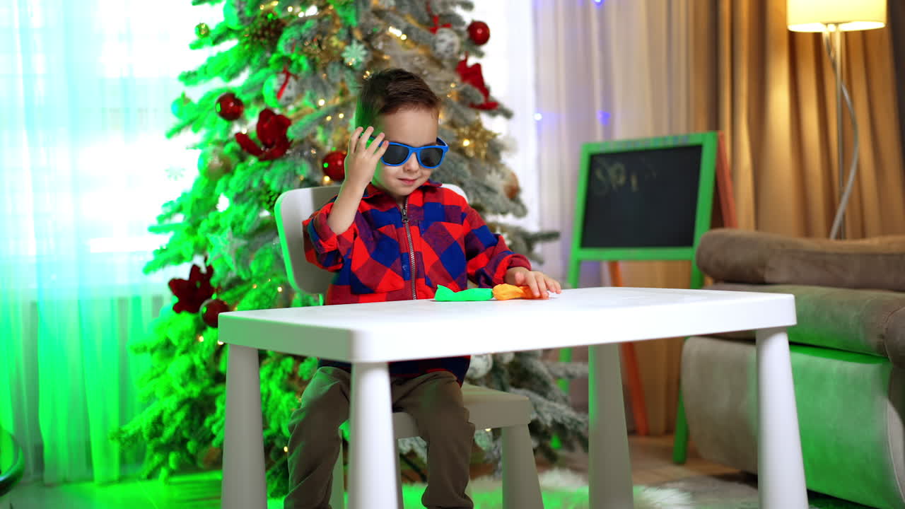 Child with sunglasses sitting at table near Christmas tree. Boy in red and blue shirt holding sunglasses at table with Christmas tree and decorations behind