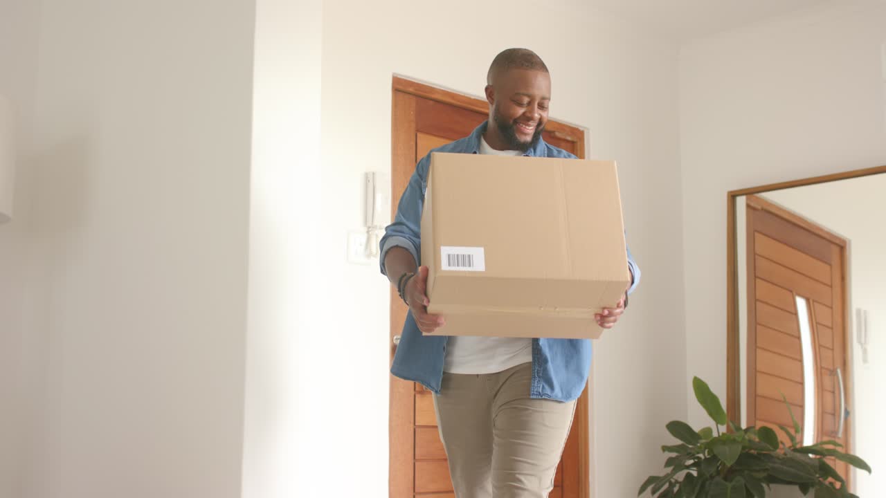African American man opening front door bringing cardboard box into living room in denim smiling