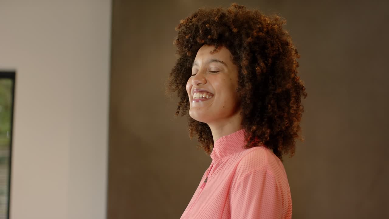 Woman in coral-pink jacket noticing camera smiling while turning profile toward front at home
