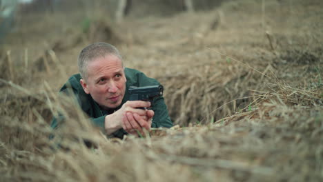 a close view of a man in a green jacket lying on the ground in a dry, grassy area while aiming a handgun