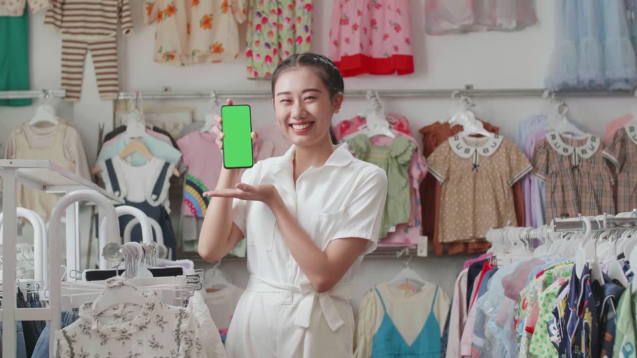 madre asiática sonriendo y mostrando un teléfono inteligente de pantalla verde a la cámara mientras hace compras en la boutique
