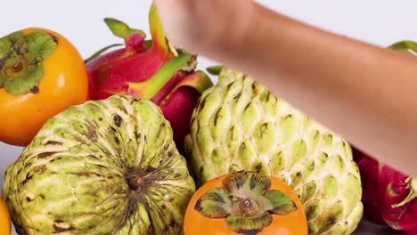 a hand reaches for and interacts with a variety of tropical fruits, including cherimoya and persimmon.
