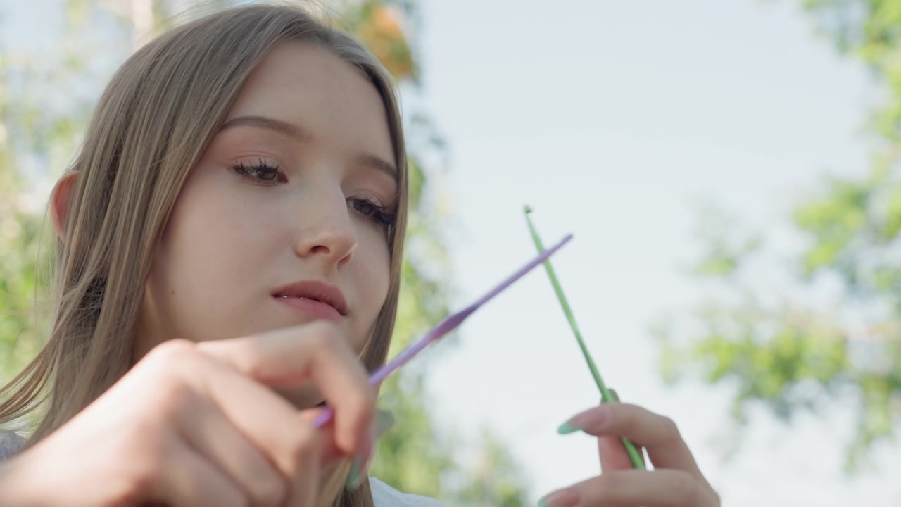 Close up of young woman demonstrating crochet technique with green and purple hooks in hand focusing on detailed finger movements. Soft natural light highlights texture of her skin