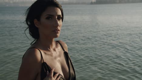 beautiful woman wearing sexy dress touches torso sensually by fingers with calm ocean in background