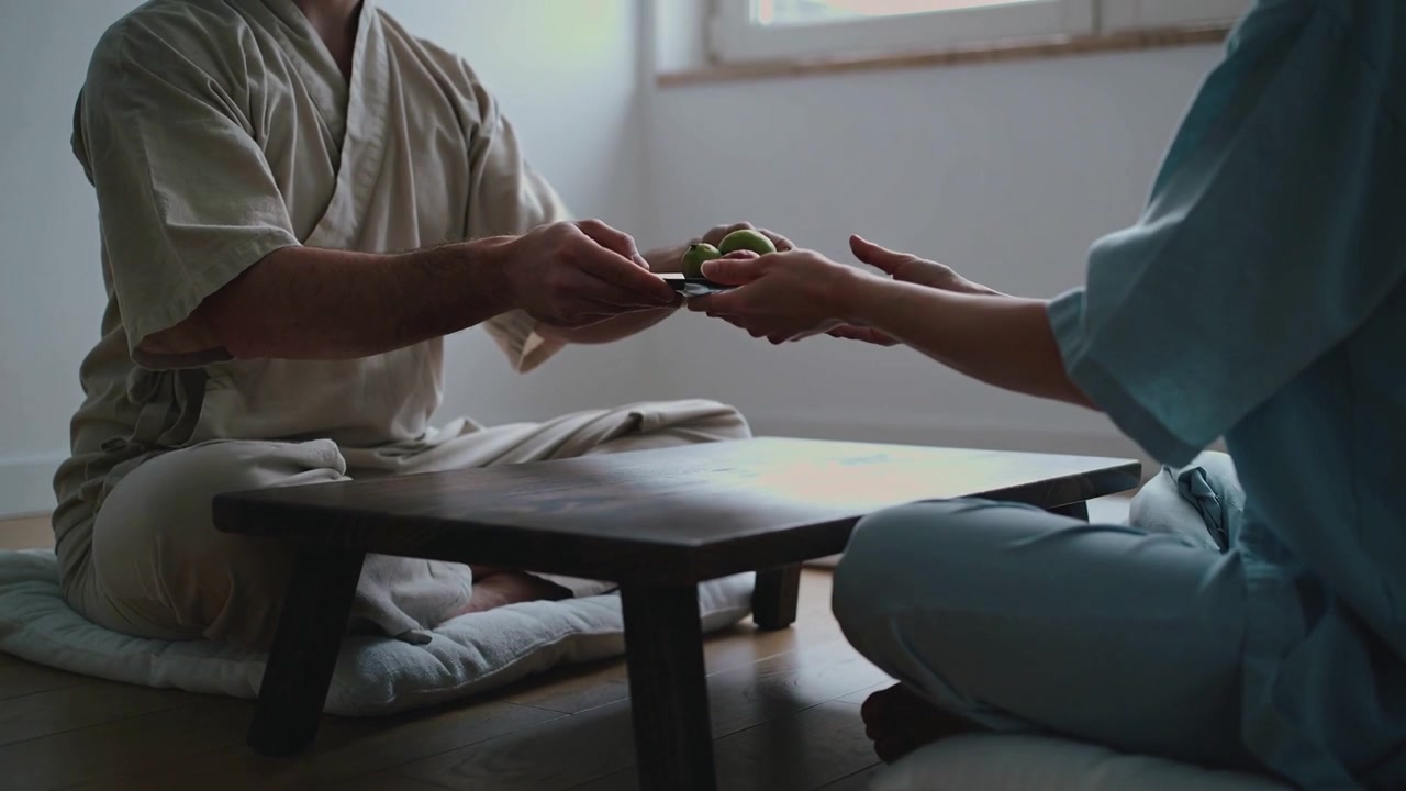 Person i kimono læner sig til venstre, rækker en tallerken til højre mod en person i blå top ved et lavt bord og deler grøn frugt.
