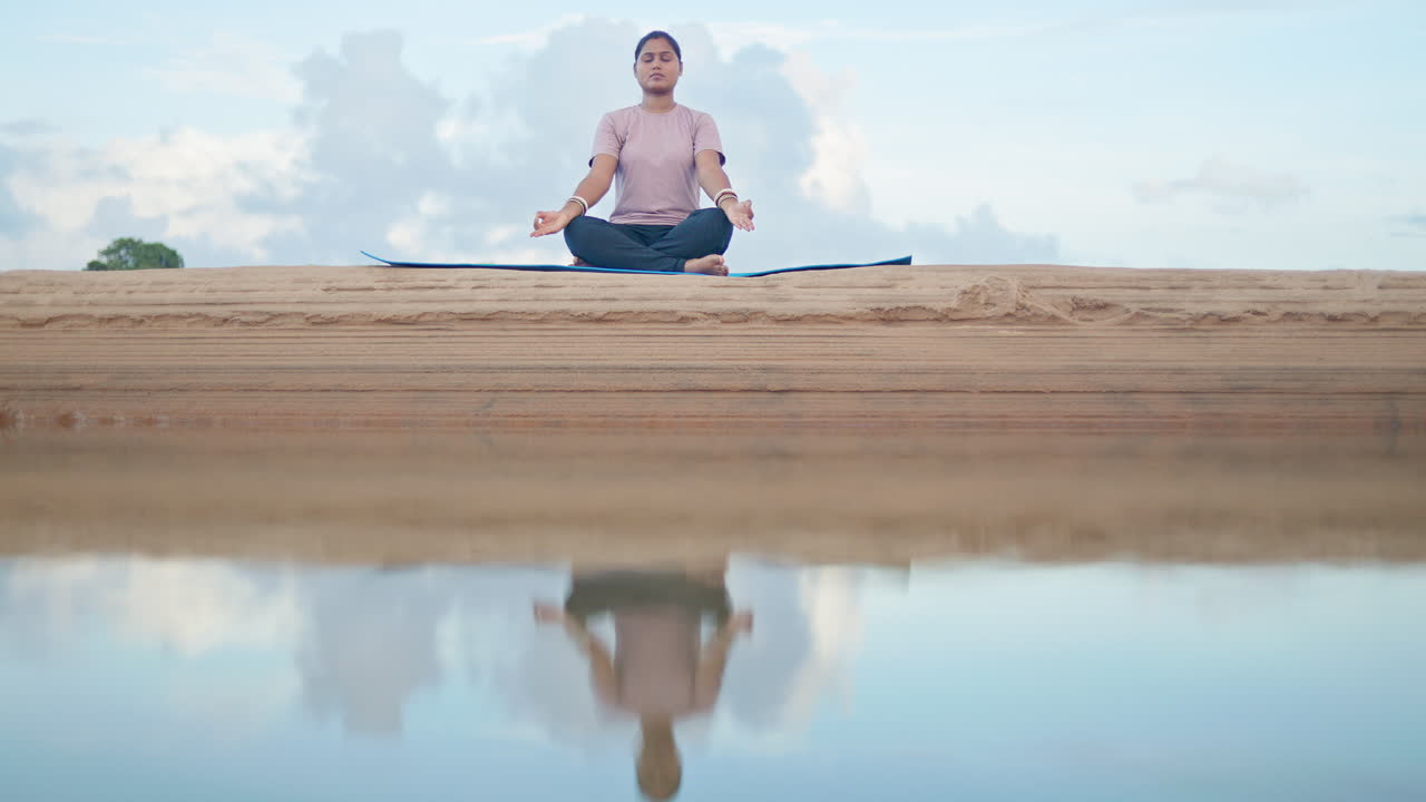 A yoga woman sitting in padmasana pose with eyes closed near a river with soft water reflection in early morning