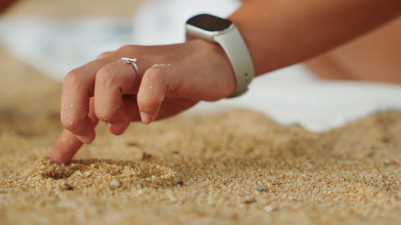 Närbild hand som ritar mönster i sanden med synlig smartklocka, resenär klottrar lekfulla spår och utforskar textur med fingertoppen, sandig ring och naglar fångar solljuset, lugn nyfiken strandstund.