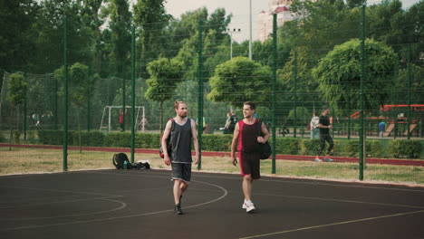 Dos-Deportistas-Guapos-Sonrientes-Caminando-Y-Hablando-Entre-Ellos,-Mientras-Salen-Juntos-De-La-Cancha-De-Baloncesto-Al-Aire-Libre-Después-De-La-Sesión-De-Entrenamiento