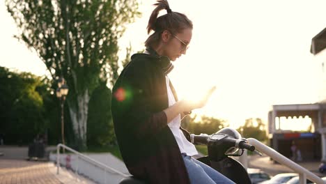 young girl in black hoodie sitting on a motorbike and typing on her mobile. sitting on moped outside in the park. son rays on