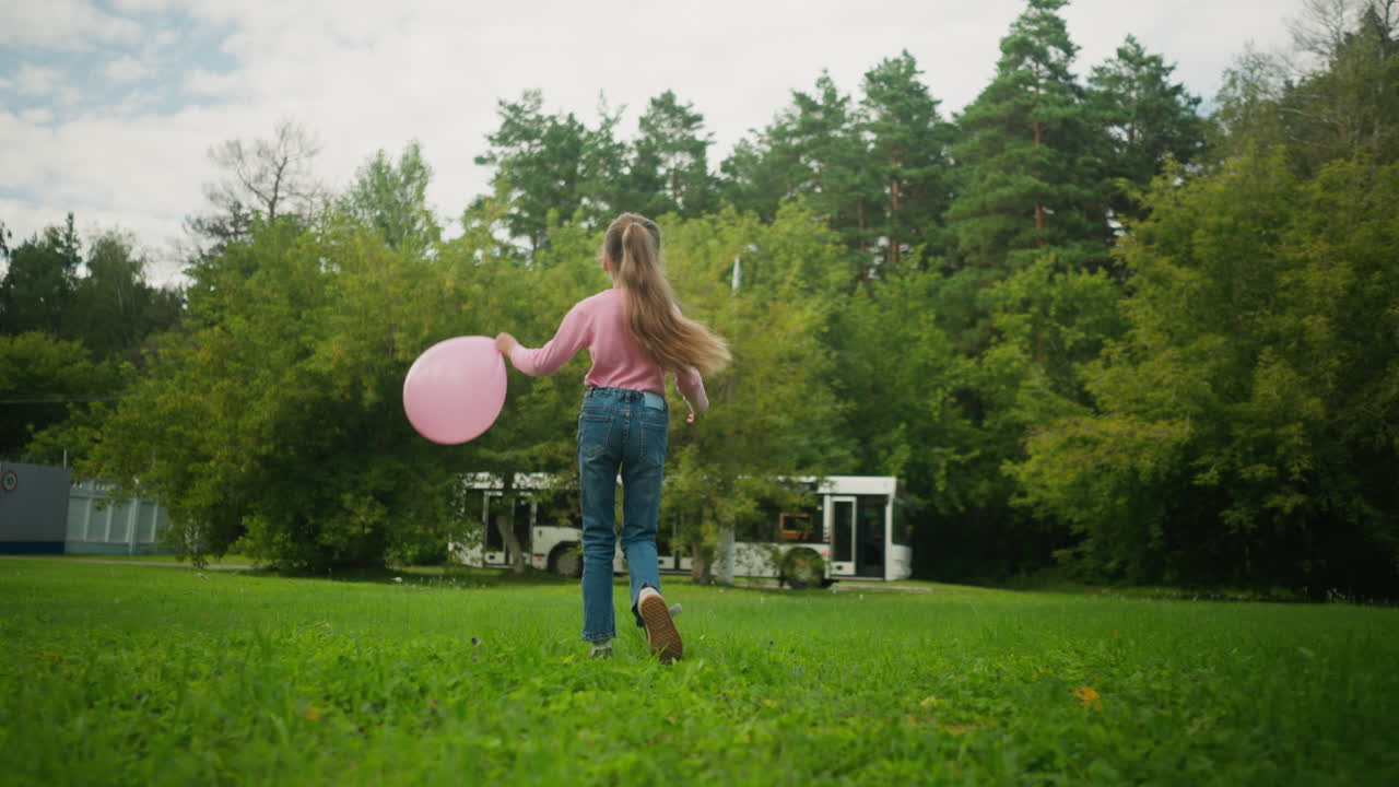 Rear view of young girl with long hair in pink top and jeans holding pink balloon while walking on green field with scattered trees, as white bus and other vehicles pass by in the distant background