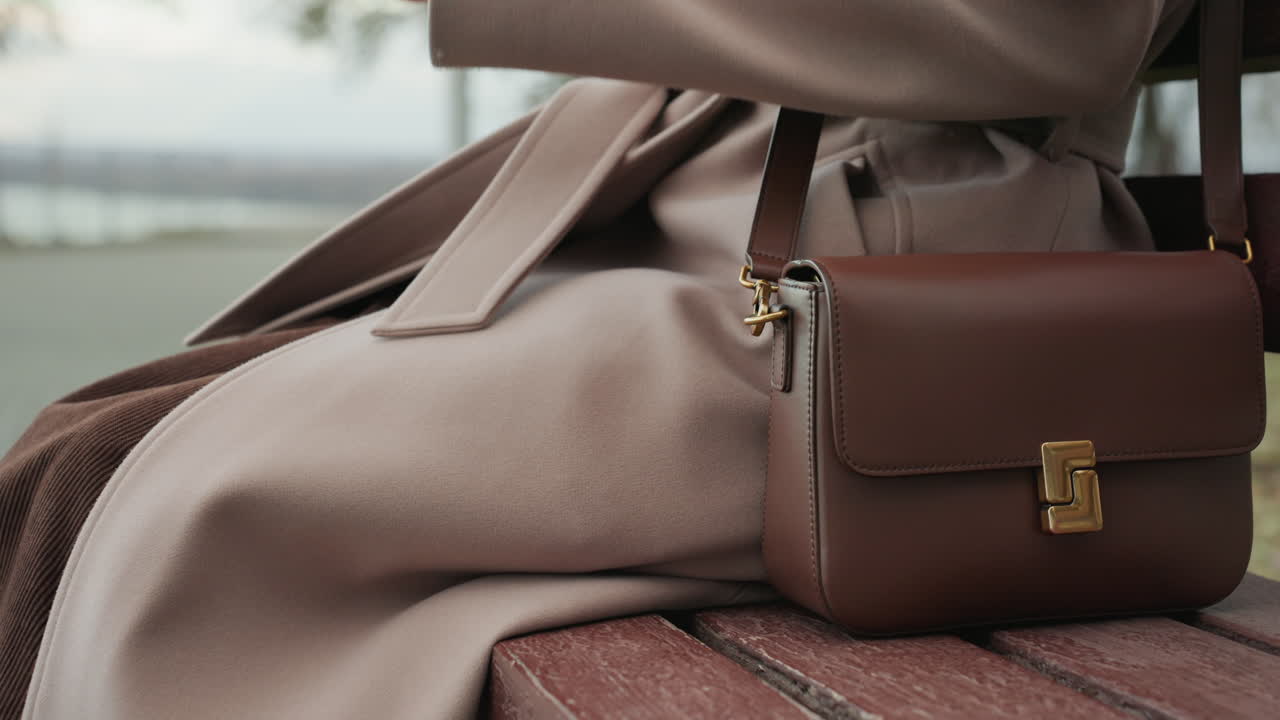 Scena in un parco lungo il fiume con una donna elegante. Una donna in stile casual poggia la mano su una borsa di pelle nel parco lungo il fiume. Pomeriggio autunnale in riva al fiume con una donna che mostra una borsa a tracolla in pelle alla moda.
