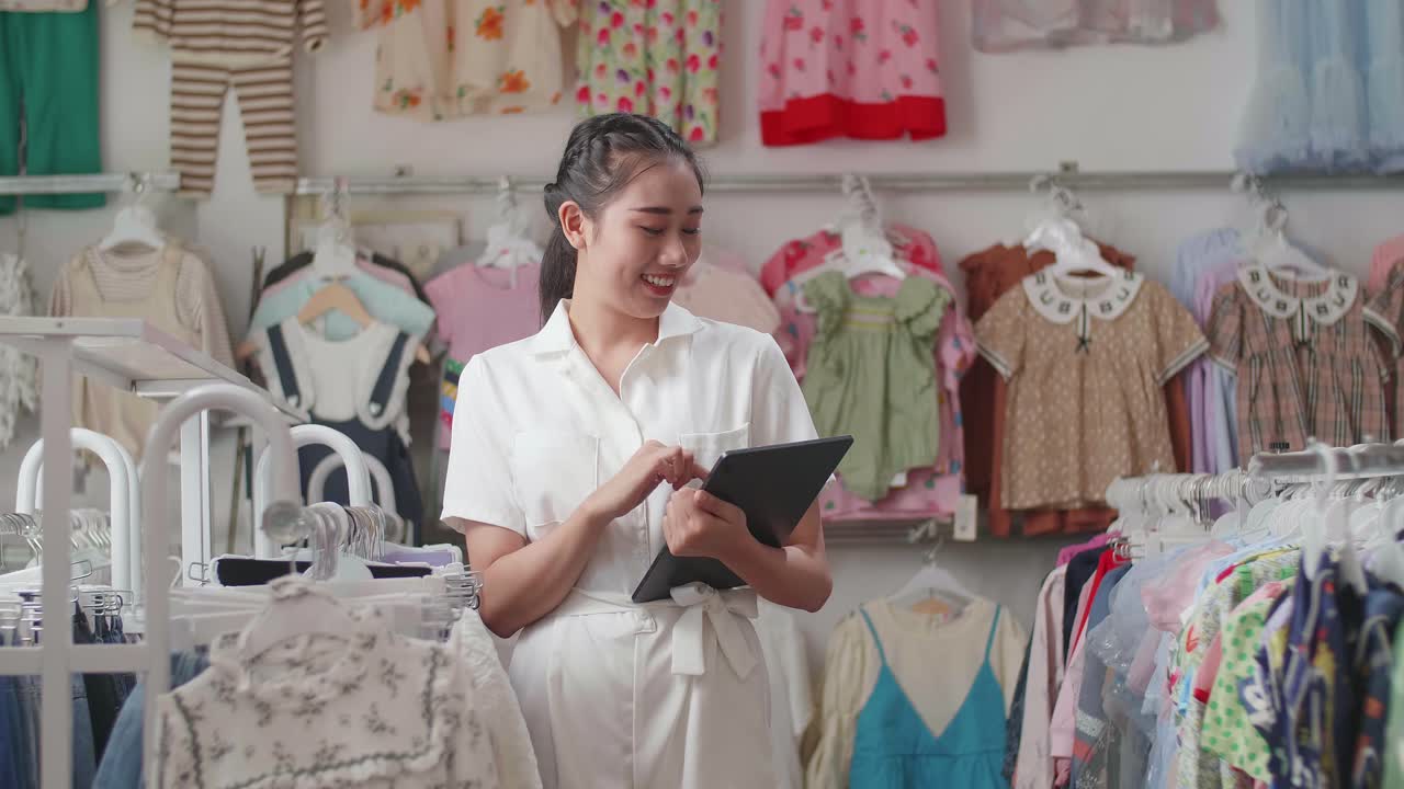 la propietaria asiática de una tienda de ropa para niños disfruta usando una tableta digital y sonriendo a la cámara mientras camina comprobando las existencias en los rieles