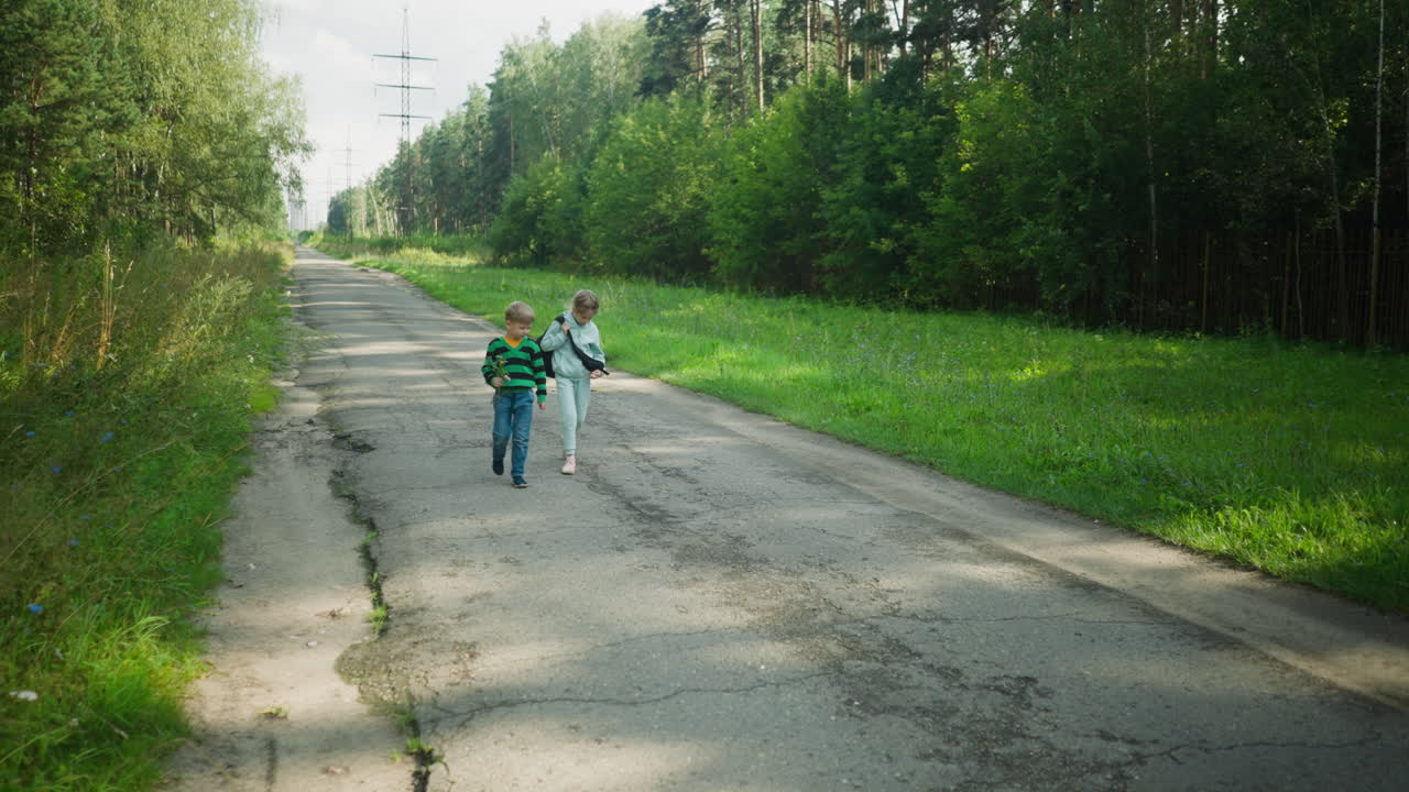 Young boy and girl walking side by side on cracked rural road surrounded by green vegetation and trees, carrying school bag and flowers in hand, calmly heading home in peaceful daylight setting