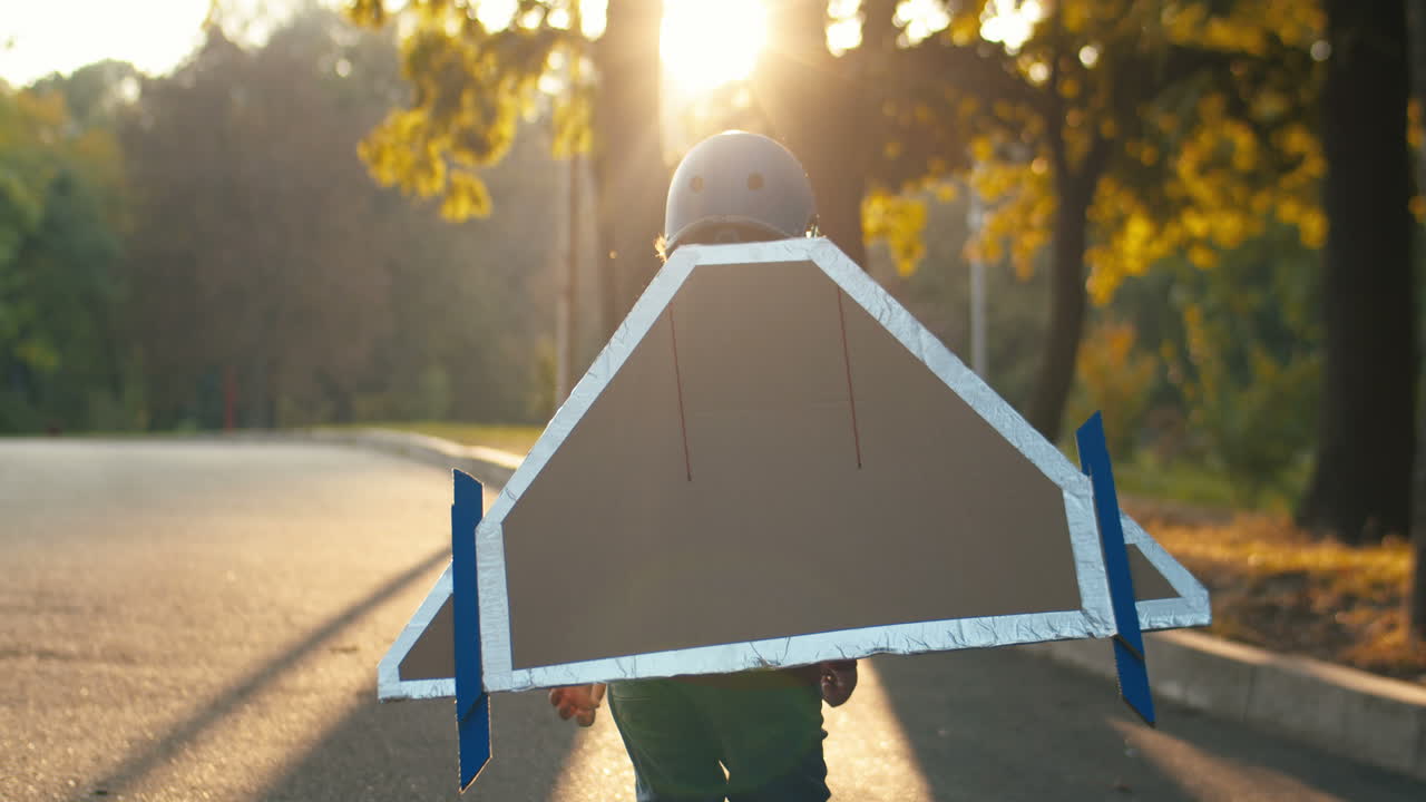 화창한 날 공원에서 달리고 조종사로 노는 마분지 비행기 날개를 가진 헬멧과 빨간 스웨터를 입은 어린 소년의 뒷모습