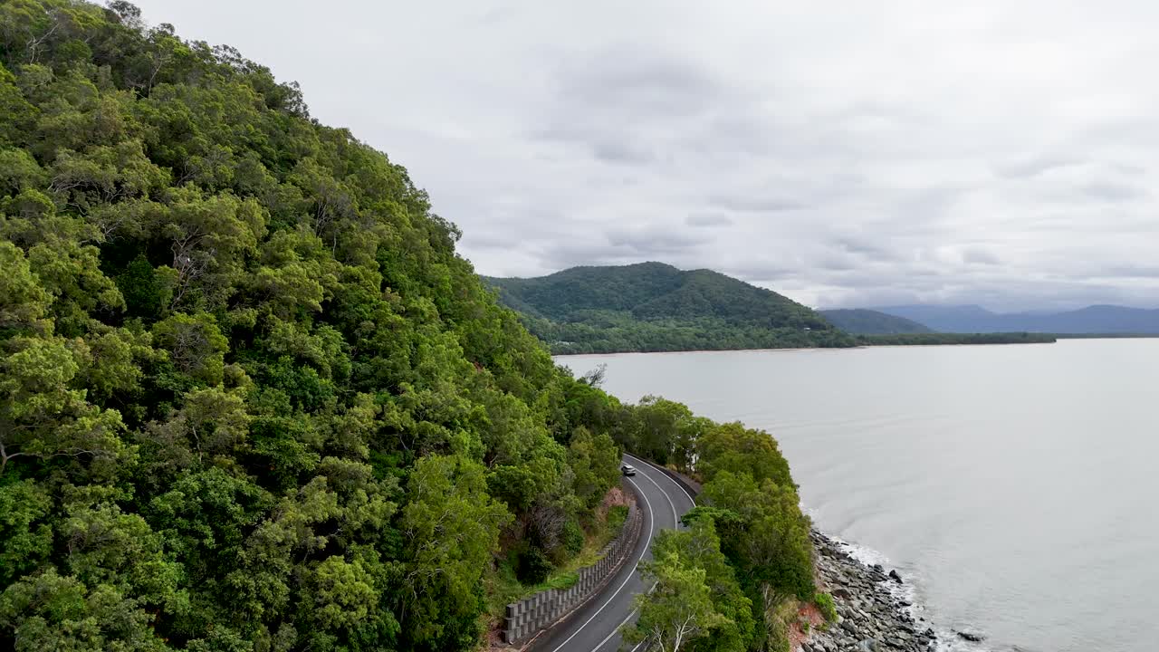Aerial view of a winding road along a lush, forested coastline with rocky shores under overcast skies