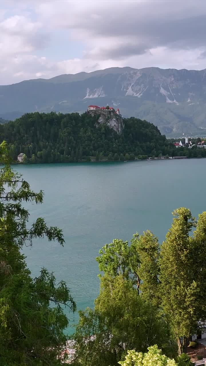 Lago e Castello di Bled, volo laterale del drone oltre gli alberi, giornata estiva nuvolosa, verticale
