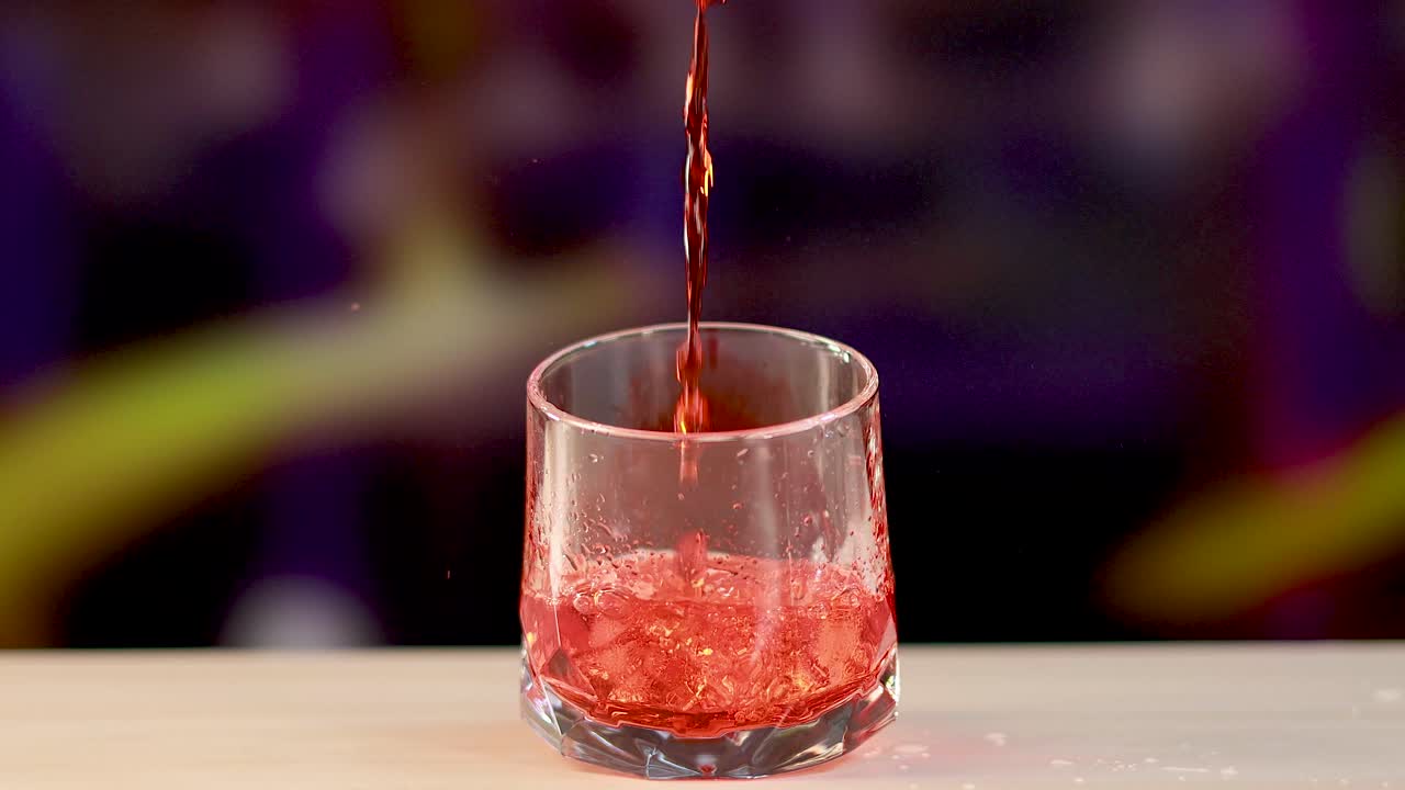 Un cocktail Negroni viene preparato versando liquido rosso su ghiaccio in un bicchiere, su uno sfondo colorato