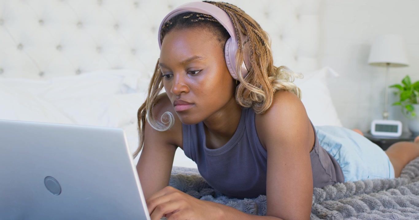 Donna afroamericana sdraiata sul letto con laptop e cuffie, che guarda lo schermo e digita per studiare.